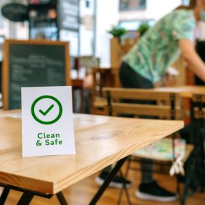 Clean and Safe sign placed on a table with waitress cleaning in the background